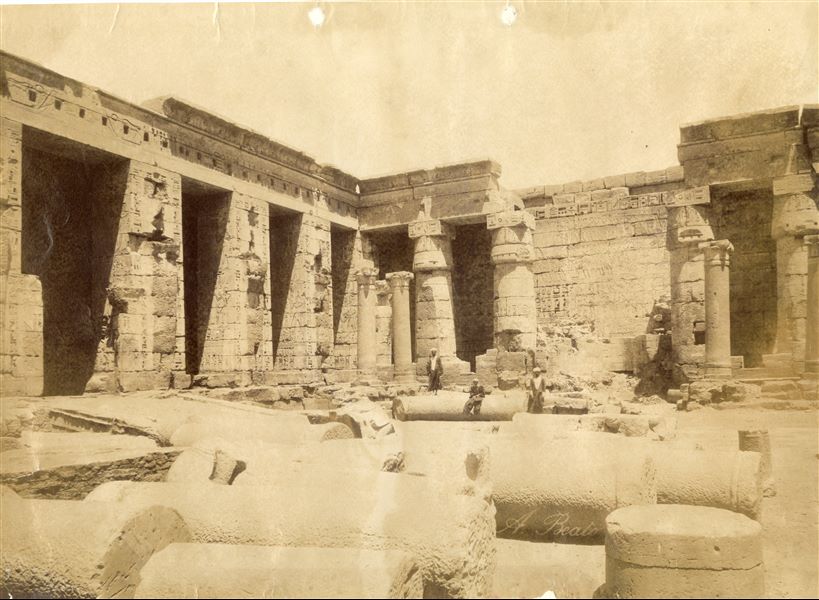 The photograph shows a view of the north-eastern corner of the second courtyard of the Temple of Medinet Habu in West Thebes, built by Ramesses III, with three locals posing for the camera between the columns, which are cut and still on the ground in the middle of the courtyard before it was cleared. The author's signature is at the bottom right. 