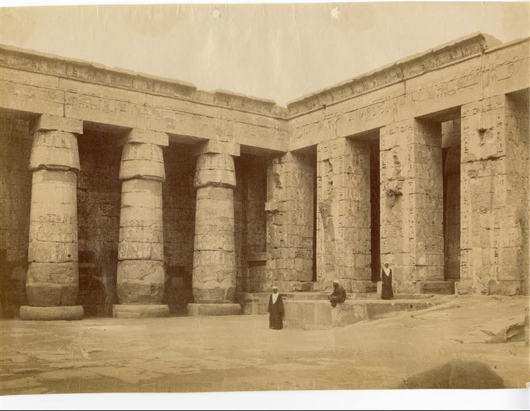 La fotografia riprende l'angolo sud-orientale del secondo cortile del tempio di Medinet Habu, costruito dal faraone Ramesse III, con tre abitanti del luogo che guardano verso l’obiettivo. La firma dell'autore è posta, a specchio, in basso a sinistra. 