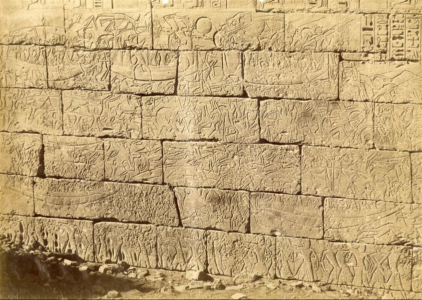 La fotografia mostra un dettaglio della battaglia navale svoltasi tra le truppe di Ramesse III e i Popoli del mare. Il bassorilievo si trova sulla parete esterna del muro settentrionale del tempio di Medinet Habu. In basso è visibile la firma dell’autore, a rovescio. 