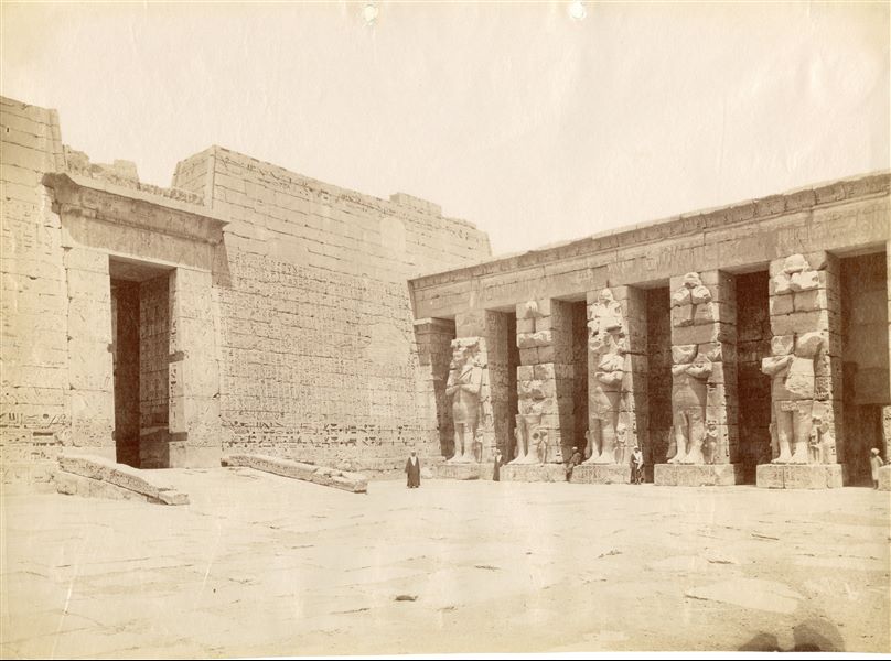 View of the northwest corner of the first courtyard in The Temple of Medinet Habu on the west bank of Thebes built by Ramesses III, and the front façade of the second pylon. The photograph shows the signature of the author, in the lower right corner in a mirror-like way and is very illegible. 
