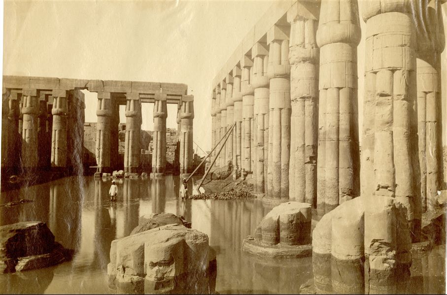 La fotografia mostra il panorama delle rovine del colonnato del cortile anteriore di Amenhotep III nel complesso templare di Amon a Luxor, inondato durante una piena del Nilo. Due Egiziani posano per il fotografo nell'acqua.  