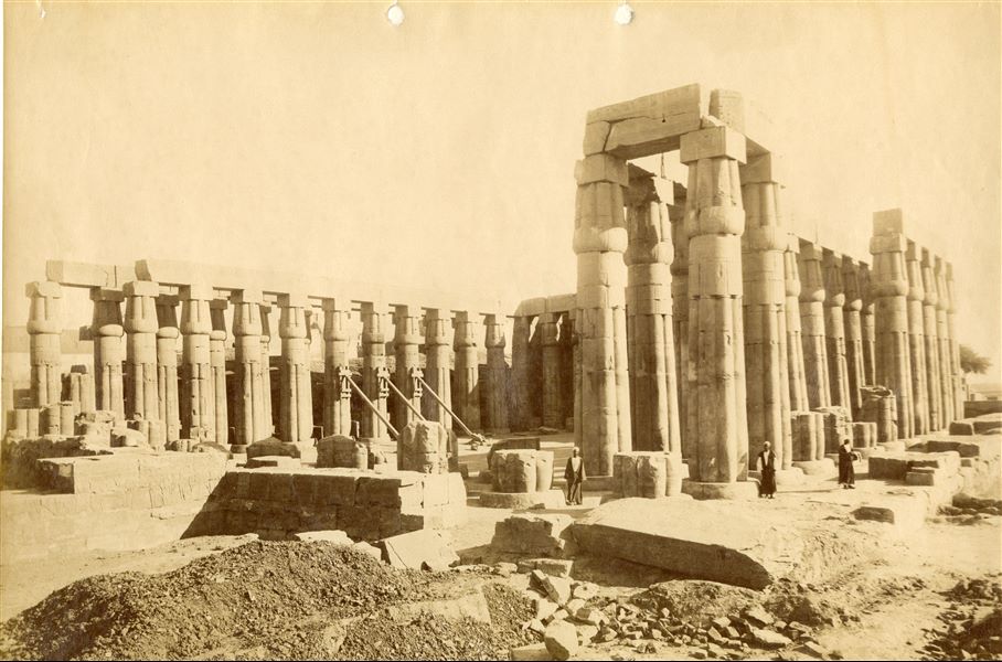 North view from the columned front courtyard of Amenhotep III in the Temple of Luxor, cleared of debris. Note three shored columns waiting to be reinforced to prevent them from falling. Three Egyptians from a distance look towards the photographer, who can be identified as Antonio Beato.