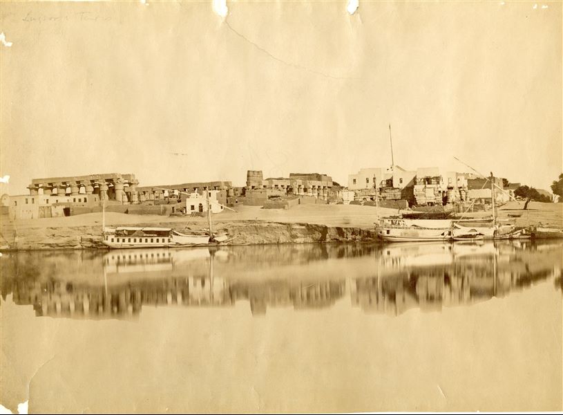 View of the Temple of Luxor photographed from the west bank of the Nile. In the foreground some feluccas (the typical boats for sailing on the river) can be made seen, and the light-coloured building on the right is the convent of the Franciscan friars. The pole from which the Italian flag must have hung is easy to locate however, in this shot barely visible. The white building in the centre was chosen by Antonio Beato as his own workshop for developing his photographs.