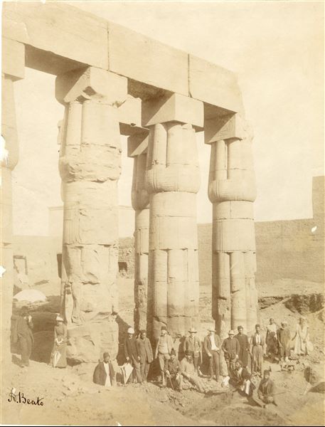 Lo scatto ritrae una schiera di personaggi (non identificati) in abiti occidentali e locali in posa per il fotografo davanti alle colonne del cortile di Amenhotep III nel tempio di Amon a Luxor. In basso a sinistra la firma dell’autore.  