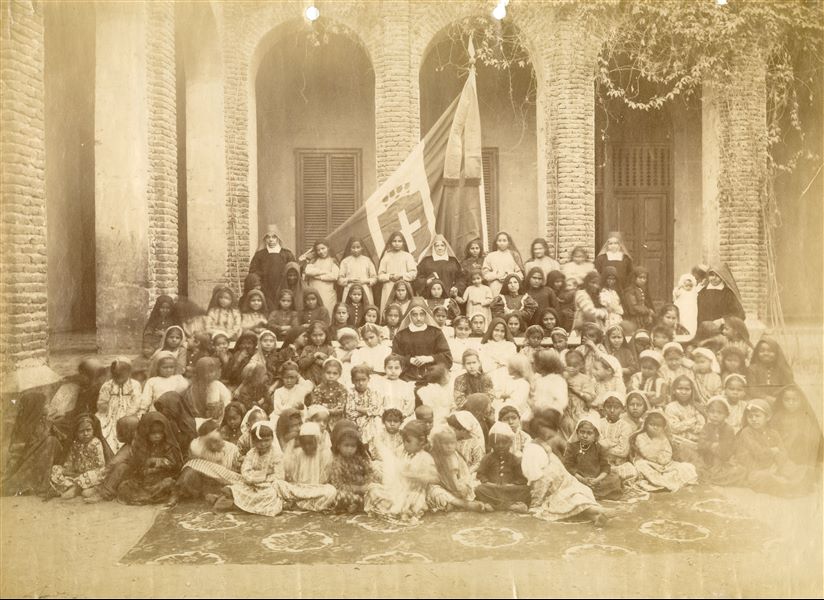 Fotografia di gruppo della scuola missionaria femminile di Luxor, nel cortile della scuola. Oltre alle suore e alle bambine, è ben visibile al centro la bandiera del Regno d’Italia. Qualche decennio dopo questo scatto, il giornale di Virginio Rosa, che scavava nel 1911 nella vicina Gebelein, riporta di una gita effettuata dalle suore e dalle orfanelle della scuola missionaria di Luxor volta a visitare gli scavi italiani, di cui è sopravvissuta anche una fotografia. 
