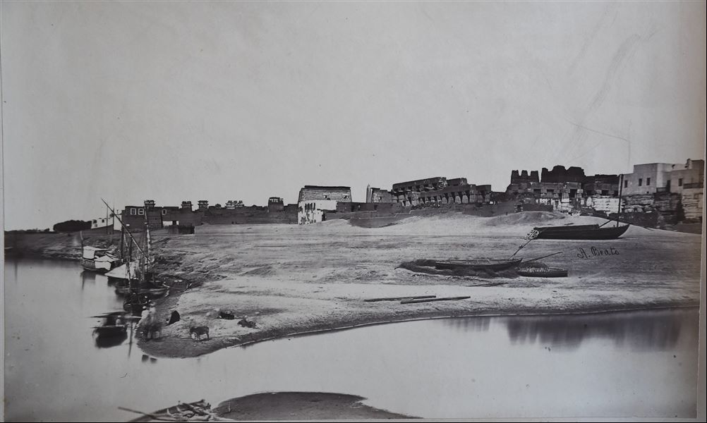 Veduta del tempio di Luxor fotografato dal Nilo, tra cui spiccano, al centro, il pilone e l’obelisco eretti da Ramesse II. Si notano alcune feluche ormeggiate, e alcuni animali andati ad abbeverarsi. La firma dell’autore si trova al centro.