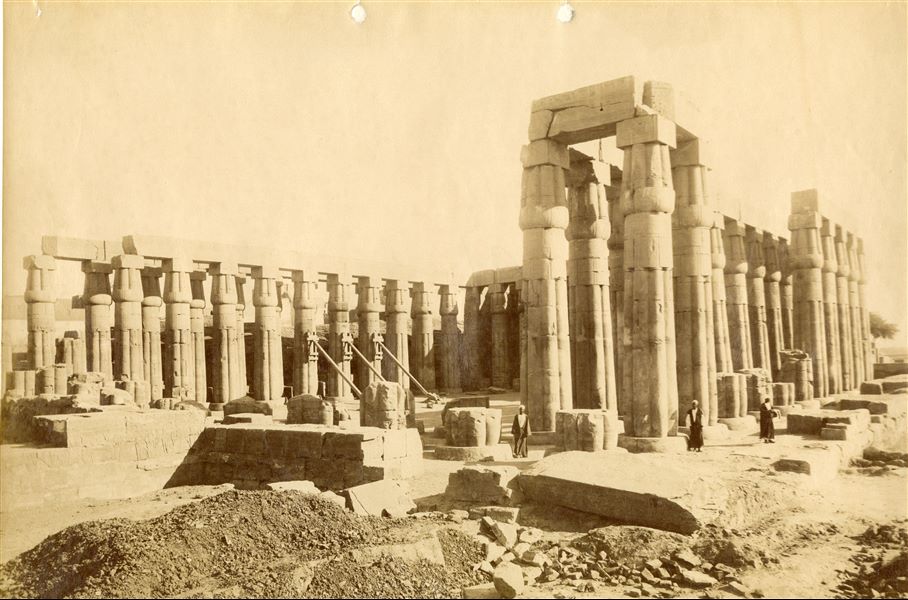 Veduta da nord del cortile anteriore colonnato di Amenhotep III nel tempio di Amon a Luxor, ripulito dai detriti. Da notare tre colonne puntellate in attesa di essere consolidate per evitare la caduta. Tre egiziani da lontano guardano verso il fotografo, che può essere identificato con Antonio Beato.