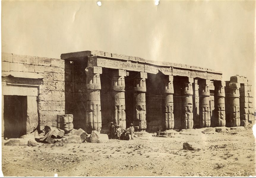 La fotografia mostra una veduta del colonnato e dell’ingresso del Tempio del faraone Seti I a Qurna, davanti a cui posano alcuni egiziani. L'opera è firmata in basso a destra. 
