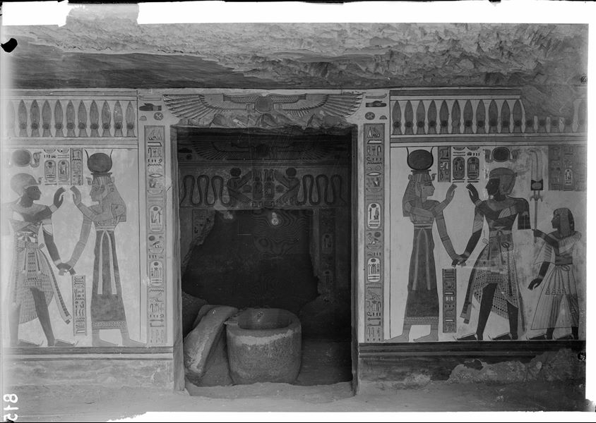 Interior of the tomb of Prince Amonherkhepeshef (QV55), at the time of its discovery. In the foreground, the prince’s granite sarcophagus in his burial chamber. On the sides, wall scenes on the southern wall: Ramesses III with the goddess Isis on the left and with the goddess Hathor on the right. Prince Amonherkhepeshef is also included in both scenes. Schiaparelli excavations. (Original label: wall in the first room, opposite the entrance and the door leading to the sarcophagus chamber). 