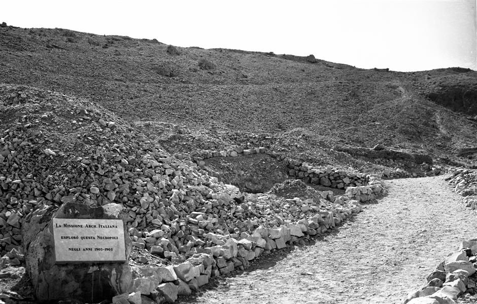 Veduta del versante sud della Valle delle Regine. In primo piano, la targa commemorativa degli scavi italiani nella valle, dietro la targa è visibile l’ingresso, delimitato da un muretto di pietre, alla tomba della principessa Ahmose (QV47). Scavi Schiaparelli. 