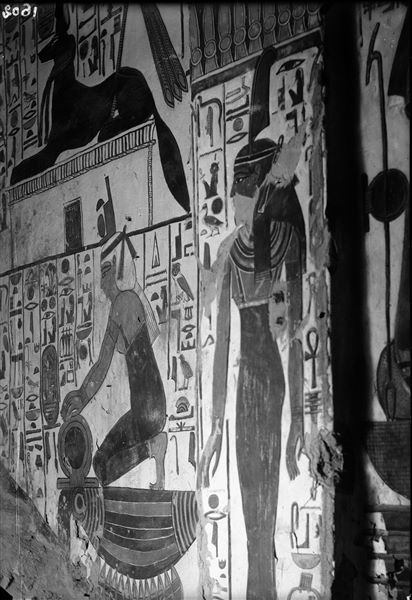 Corridoio discendente, parete ovest, scena 26. A destra, la dea Maat (scena 28, rappresentata nel lato sinistro dell’accesso alla camera funeraria), a sinistra, inginocchiata, la dea Iside. Nel registro superiore, il dio Anubi nella sua forma animale, sopra un santuario. Le iscrizioni riportate sono espressioni benevoli enunciate dalle due divinità nei confronti di Nefertari. 