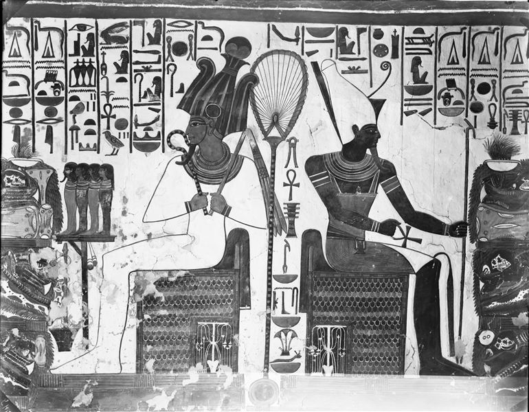 Primo annesso orientale (alcova), parete est, scena 18. Rappresentazione della zona centrale della parete, dove sono assise due divinità, da una parte Osiride, dall’altra Atum. Essi stanno simmetricamente ricevendo un tavolo colmo di offerte da Nefertari, le cui due rappresentazioni non sono qui visibili.