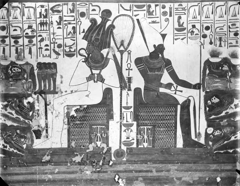 Primo annesso orientale (alcova), parete est, scena 18. Rappresentazione della zona centrale della parete, dove sono assise due divinità, da una parte Osiride, dall’altra Atum. Essi stanno simmetricamente ricevendo un tavolo colmo di offerte di offerte da Nefertari, le cui due rappresentazioni non sono qui visibili.