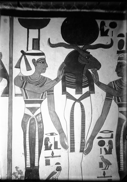 Primo annesso orientale (alcova), parete ovest, lato sud, scena 15. Una divinità mummiforme con testa di ariete e disco solare, che richiama alle caratteristiche di Osiride e di Ra, è assistita dalle dee Neftys (a sinistra) e Iside (a destra).