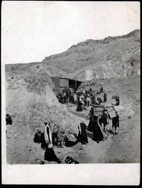 Transporting antiquities from the Italian Archaeological Mission’s camp to Luxor. The camels are about to leave the camp, where a brick shelter is also visible. One of the figures in front of the shelter is Bolos Ghattas, the mission's dragoman (guide and interpreter). In the background, the coffins found at the time of the discovery of tombs of Sethiherkepeshef (QV 43) and of Khaemwaset (QV 44) are visible.