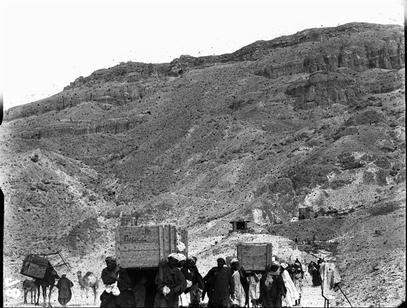 Transporting crates from the camp in the Valley of the Queens (in the background) to the Nile during the final stages of the excavation. Notable on the right is a camera covered by cloth. Schiaparelli excavations.
