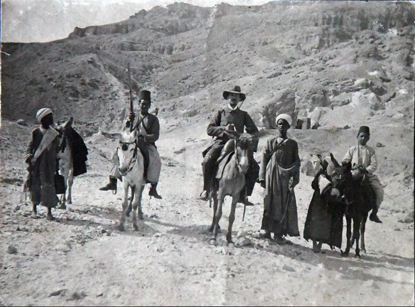 Photograph taken in the Valley of the Queens, during excavations in 1903. In the centre is Francesco Ballerini, while to his right holding a rifle, is the dragoman (guide and interpreter) Bolos Ghattas. In the background, to the right, the ruins of the Coptic monastery of Deir Rumi. Angelo Sesana Archive. 