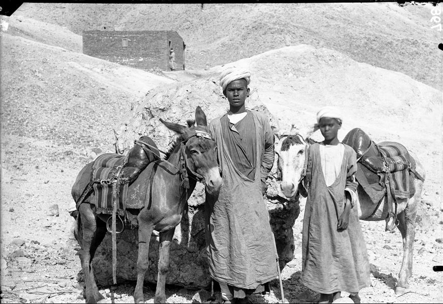 Primo piano di due giovani membri della missione italiana, insieme a due muli. Sullo sfondo, la struttura in mattoni costruita per la missione archeologica. Scavi Schiaparelli. (Didascalia originaria: asinai del vicino villaggio di Gurnah addetti al servizio della Missione Archeologica).