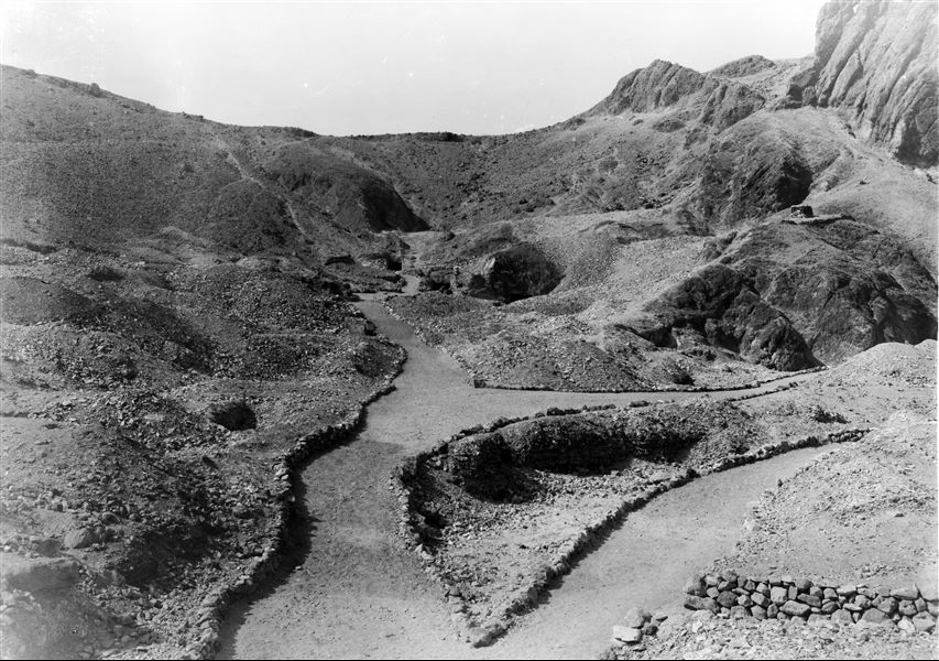 View of the last section of the Valley of the Queens. Visible from right to left are the entrances to tombs QV44 (of Prince Khaemwaset) and QV43 (of Prince Setiherkhepeshef). Along the path, the plaque commemorating the Italian excavations in the Valley can be seen, followed by the barely visible shaft of the tomb of Princess Ahmose (QV47). Schiaparelli excavations.
