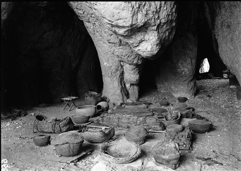 Inside the large natural caves. Given the more moderate temperature inside, they were used for preliminary study and examination of the excavated materials. On the folding stool there are brushes used for dusting the objects. The image displays numerous deceased gathered together, mainly in baskets and wrapped in mats. Schiaparelli excavations. 