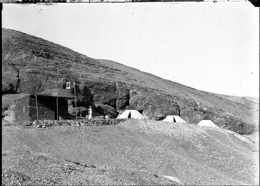 Camp of the Archaeological Mission. In the foreground, there is a small more recently built construction, as can be inferred due to the dark colour of the mud mortar. This type of construction was built on almost all excavation sites and some of them are still visible. The camp was always accompanied by the Italian flag. In the centre is the figure of the gafir (local guard). Schiaparelli excavations. 