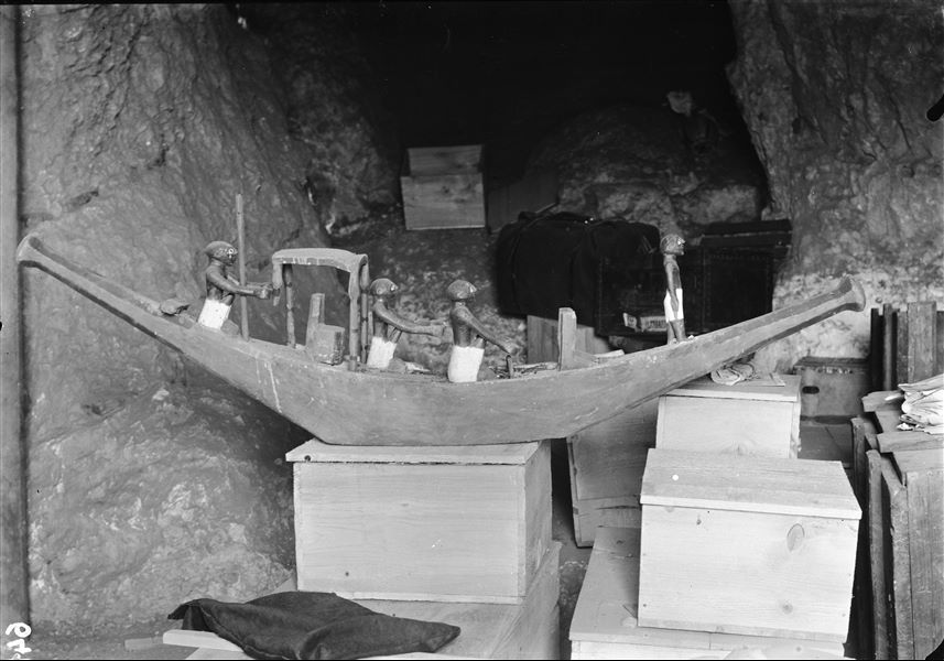 Tomb of Ini. Before being packed for transport, the objects were inventoried and photographed, often gathered in groups. The picture shows the preparation of packing special crates to house the materials. In the background there are bags and trunks. In the foreground, a model of one of the two boats with a captain, helmsman and rowers (S.13272). Schiaparelli excavations. 