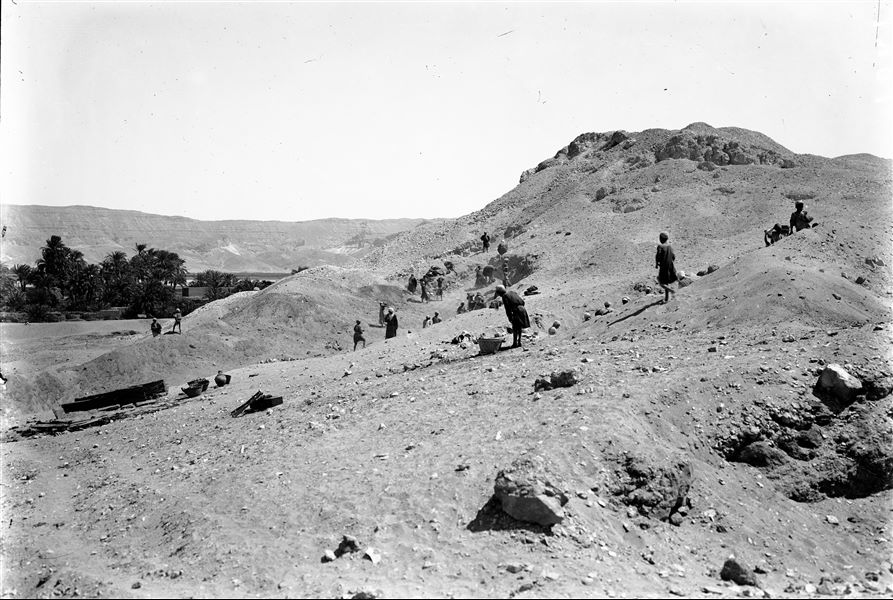 Excavations north of the hill, along the slopes. On the left, fragmentary remains of wooden sarcophagi. Further towards the centre of the image, is a box containing photographic materials and a foldable chair. Farina excavations. 