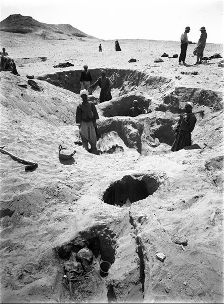 Northern necropolis. Burial pits, human remains with grave goods beside them. In the grave in the centre, the deceased is still wrapped in matting. On the left, there is a hoe and a basket, used to remove excavated material. On the right, Giovanni Marro can be seen holding an artefact in his hands, with Giulio Farina next to him. Farina excavations. 