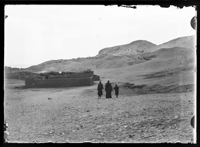  Two adult Egyptians and a child in front of a modern mud-brick fence, photographed during excavations, which can be seen in the background, on the northern hill at Gebelein. Presumably Schiaparelli excavations.