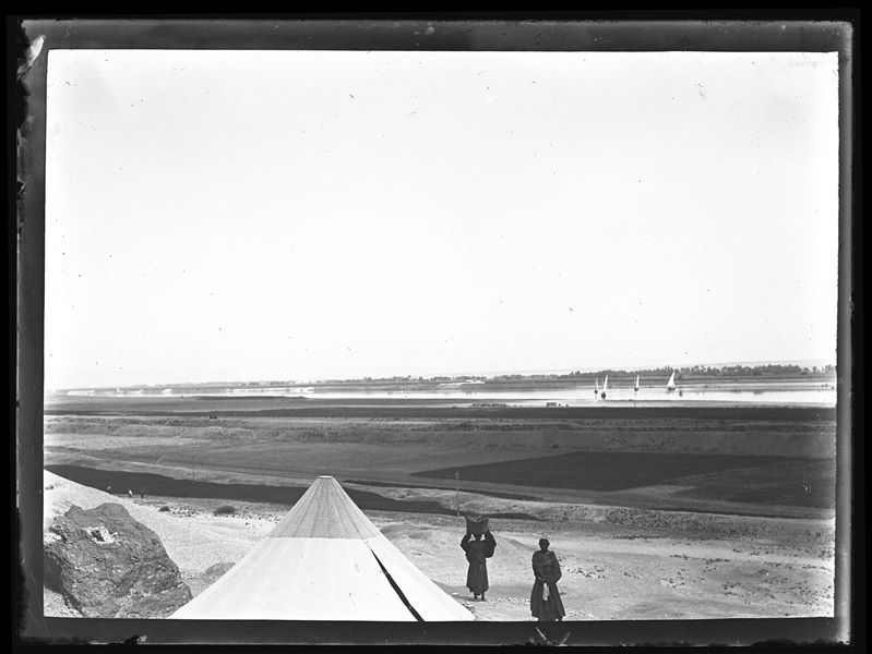  At the bottom towards the left, two Egyptians can be seen next to a tent belonging to the Italian Archaeological Mission, in Gebelein. In the background, some boats sailing on the Nile River. Schiaparelli excavations.