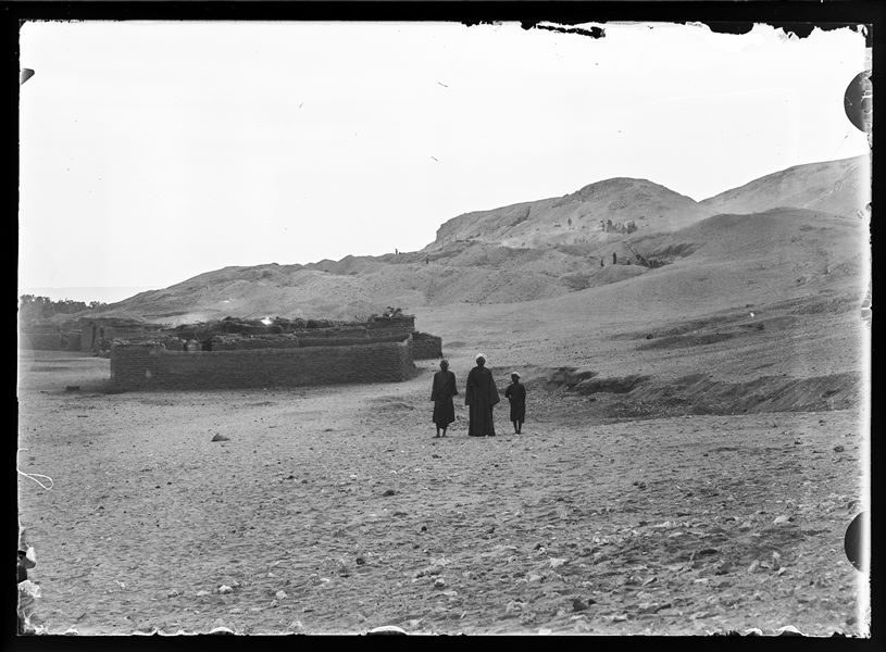  Two adult Egyptians and a child in front of a modern mud-brick fence, photographed during excavations, which can be seen in the background, on the northern hill at Gebelein. Presumably Schiaparelli excavations.
