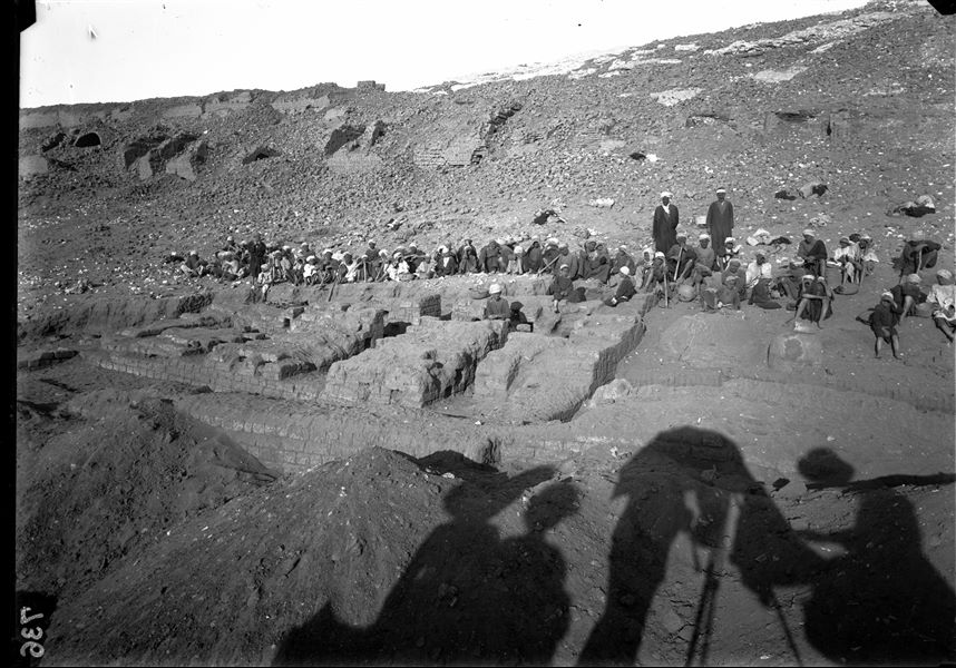 Excavations at the foot of the southern hill, western side. The workers (many of them very young) are on a break in order to photograph the site without their presence, and to avoid the inconvenience caused by dust clouds when excavating. Whilst taking the photo, the sun has projected the shadows of the people close to the camera onto the ground, including the figure of Ernesto Schiaparelli who is under a parasol. Schiaparelli excavations. 