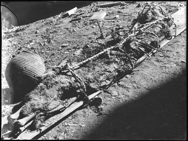 Burial lying on wooden planks, held by ropes, photographed at the moment of discovery. Some scattered grave goods can be seen around it. Schiaparelli excavations. 