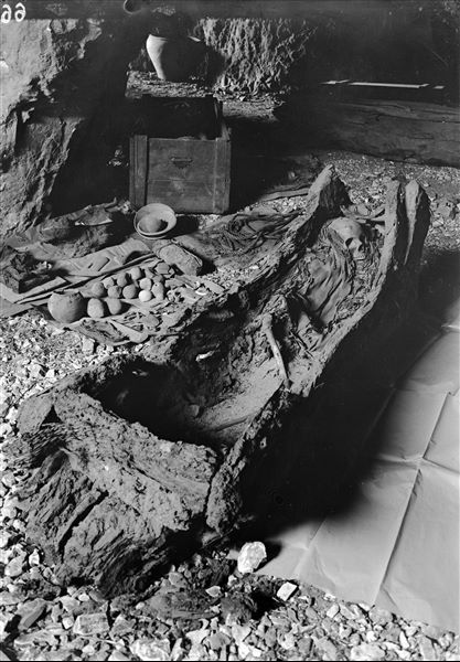 Burial in a tree-trunk coffin, lying on its side. The image was taken in the large cave used by the camp. Around the coffin, there are some grave goods, as well as those from other burials. The materials were kept separate to protect and keep them intact before they were packaged. Schiaparelli excavations. 