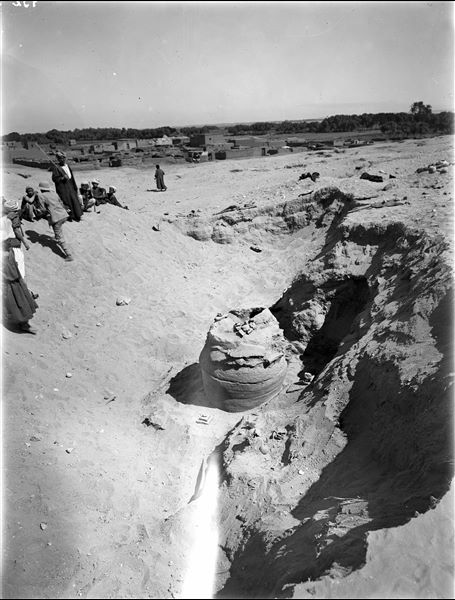 Scattered burials on the slopes of the northern hill, next to the modern village of Abu Hummas. In the foreground, a large terracotta vase made of several stacked circular ceramic pieces probably containing a deceased individual. On the left, next to the rais (chief of the workmen) with his cane raised, is Giulio Farina. Farina excavations. 