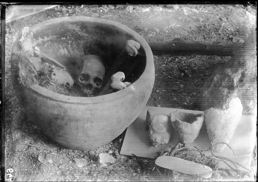 ​Scattered burials on eastern slopes of north hill. Vase with human remains. Often, inside or outside of the burial vase, there were grave goods present. In this example, there are some fragments of very simple conical vases probably from predynastic times, and the remains of a vegetable fibre net. Schiaparelli excavations. 