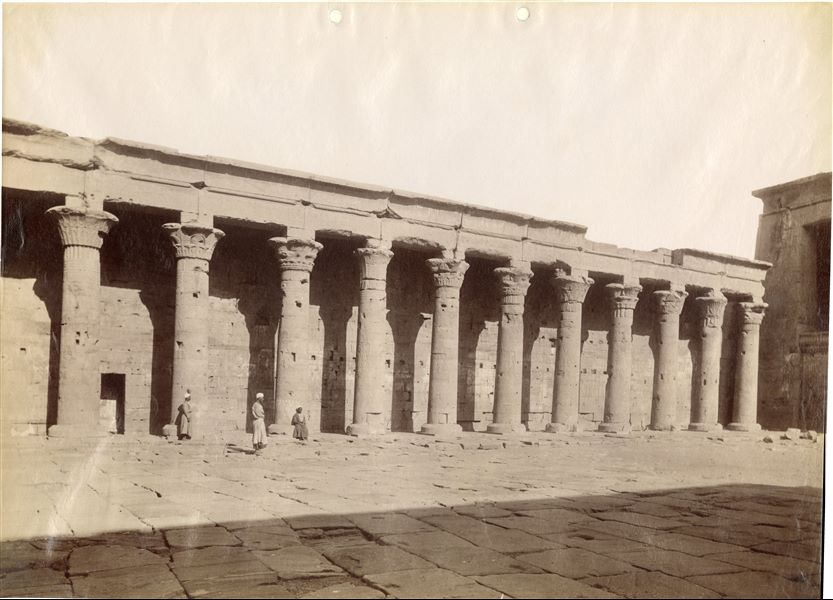The photograph (same as the previous one) shows a view of the west side of the colonnade of the courtyard in front of the entrance to the first hypostyle hall from the Temple of Horus at Edfu, on the right. Three egyptians are shown in front of the columns. Based on the calligraphic style of the caption, the shot can be attributed to Antonio Beato. 