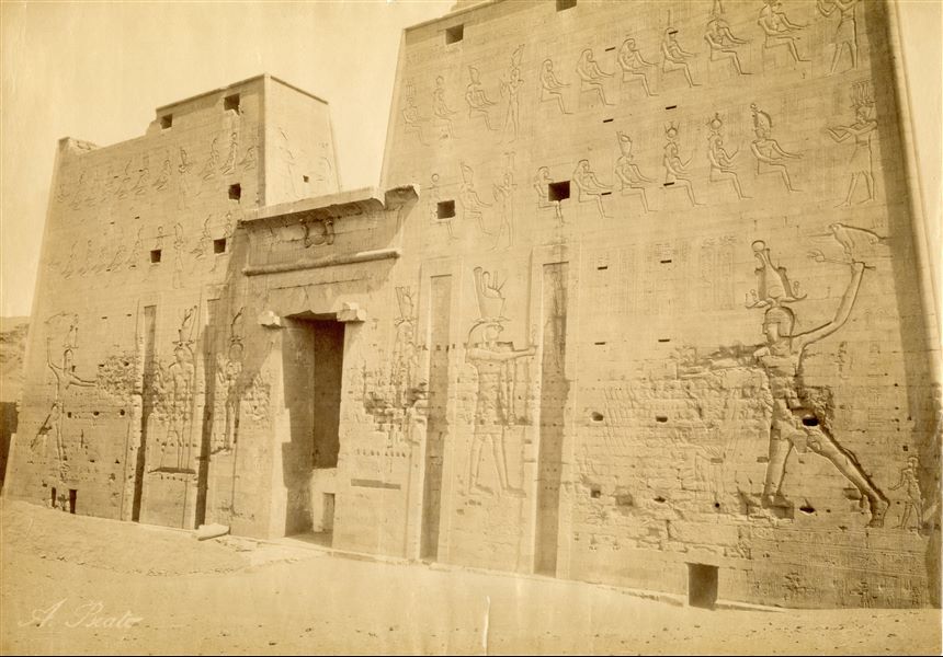 The photograph is a close-up of the entrance pylon to the Temple of Horus at Edfu, where the relief decorations with pharaohs and gods are clearly visible. It is curious to note that the monumental access is still closed by a wall with a door, built by the locals. The author's signature is visible at the bottom left. 