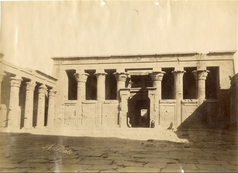 Veduta del pronao del tempio di Horus a Edfu, con due abitani locali che vi sostano davanti, e la parte occidentale del colonnato del cortile antistante. La firma dell'autore è apposta in basso a sinistra.  
