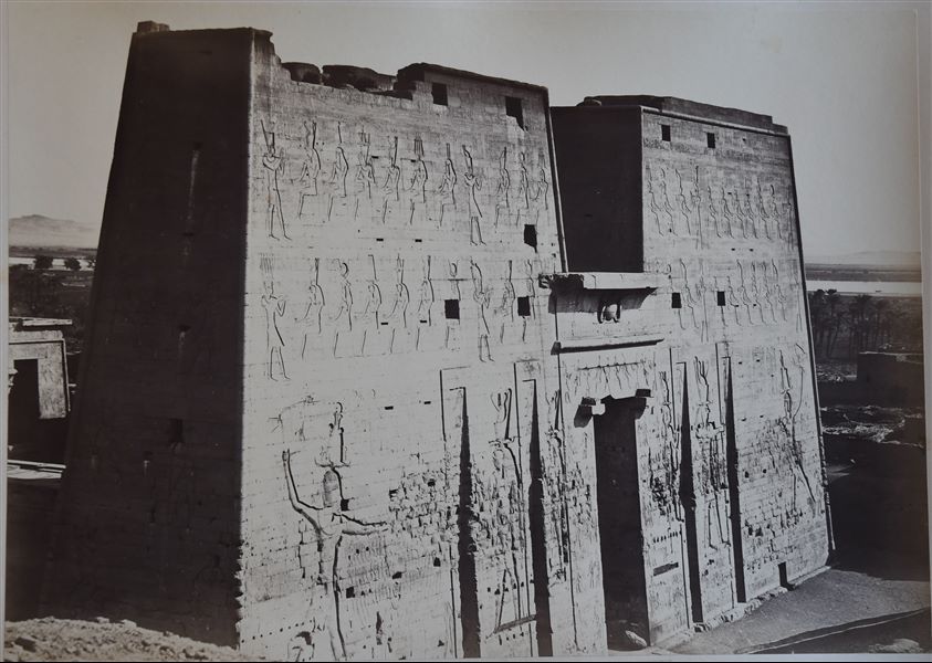 The photograph shows the front side of the first pylon that accesses the Temple of Horus at Edfu. The Nile is visible in the background. 