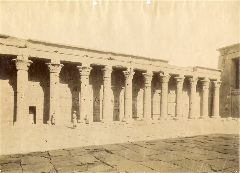 The photograph shows a view of the west side of the colonnade from the courtyard in front of the entrance to the first hypostyle hall from the Temple of Horus at Edfu (on the right). Three egyptians are shown in front of the columns. Based on the calligraphic style of the caption, the shot can be attributed to Antonio Beato. 