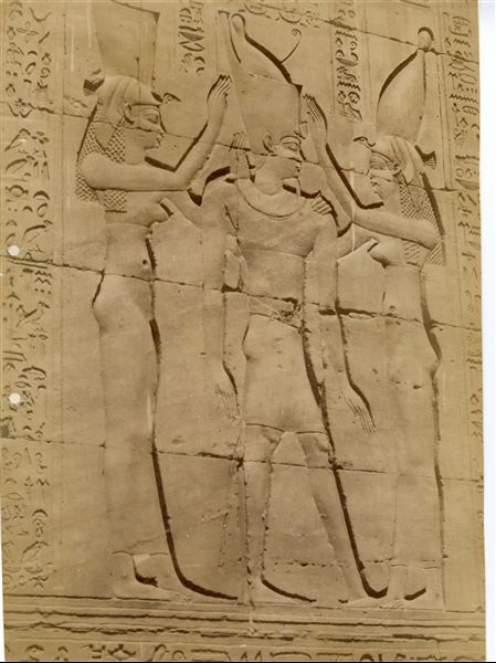 The photograph shows a relief scene from the Temple of Horus at Edfu. Pharaoh Ptolemy VI Philometor is between two female deities in a purification scene, one deity is wearing the white crown of Upper Egypt, the other the red crown of Lower Egypt. 