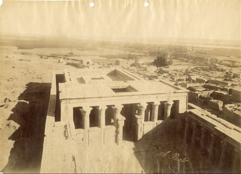 Photograph taken from the top of the entrance pylon to the courtyard and pronaos of the Temple of Horus at Edfu; where the hypostyle hall and other inner rooms can be entered. It is interesting to note that the “windows” on the ceiling of both the pronaos and the innermost part of the temple which probably gave light to the rooms, are now closed. In the background, the egyptian landscape. The author's signature is clearly visible at the bottom. 