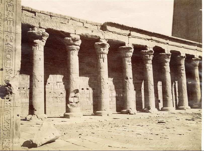 The photograph shows a view of the eastern side of the colonnade from the courtyard in front of the façade of the Temple of Horus at Edfu, with two egyptians seated at the base of the columns. On the left, part of the jamb of the pronaos is visible, while on the right one can glimpse part of the inner wall of the entrance (first) pylon to the temple. The author's signature is visible at the bottom left. 
