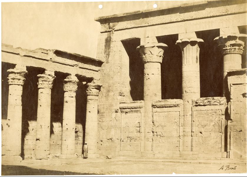 La fotografia riprende l'angolo nord-occidentale dell’imponente pronao del tempio di Horus a Edfu e parte del colonnato del cortile antistante. Un abitante del luogo posa per il fotografo davanti ad una colonna. La firma dell'autore è posta in basso a destra. 