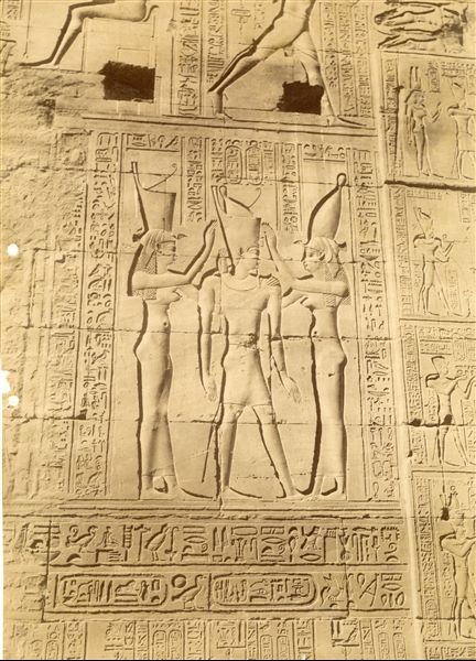 The photograph shows a sacred scene from the Temple of Horus at Edfu, Pharaoh Ptolemy VI Philometor between two female deities, one wearing the white crown of Upper Egypt, the other the red crown of Lower Egypt. 