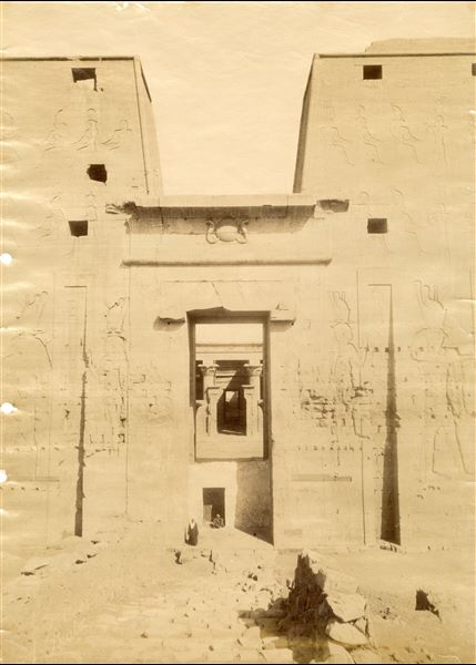 The photograph shows a close-up of the entrance pylon to the Temple of Horus at Edfu, through which there is a view of the courtyard and pronaos. It is curious to note that the monumental access is still closed by a wall with a door, built by the local inhabitants. The author's signature is faintly legible at the bottom left. 