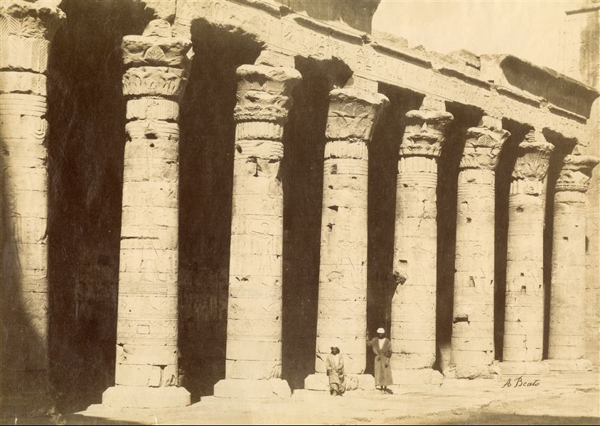 Two local inhabitants are photographed in front of the columns of the western side of the colonnade, from the courtyard in front of the hypostyle hall of the Temple of Edfu, part of the pronaos can be seen on the right. The author's signature is at the bottom right. 