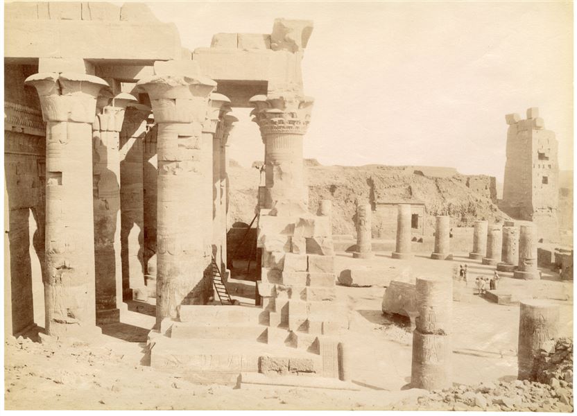Nella fotografia sono rappresentati l'atrio ipostilo esterno, costruito da Tolemeo XII, e il cortile anteriore, di Augusto, del tempio di Sobek e Haroeris a Kom Ombo visti da nord-ovest. In secondo piano, a destra, è visibile il pilone d'accesso al recinto sacro. Sulla base dello stile calligrafico della didascalia è possibile attribuire lo scatto ad Antonio Beato. 
