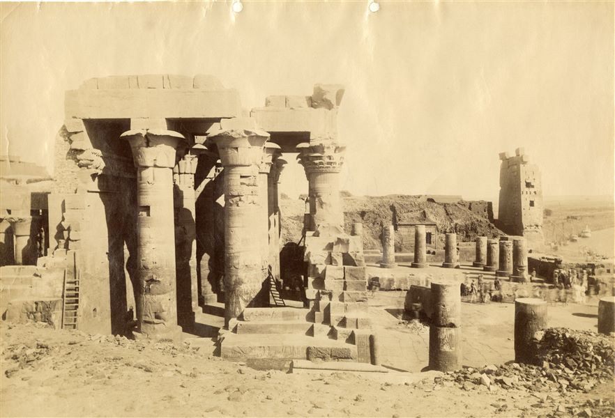 Veduta settentrionale del cortile anteriore e dell'atrio ipostilo dei resti del tempio di Sobek e Haroeris a Kom Ombo. Lo scatto, che ha avuto una lunga esposizione, mostra sulla destra diverse persone che si stanno muovendo, presumibilmente per ripulire il tempio dai detriti, risultando pertanto mosse e sfocate. Sullo sfondo a destra, il Nilo e alcune feluche, le imbarcazioni tipiche egiziane per navigare lungo il fiume. 